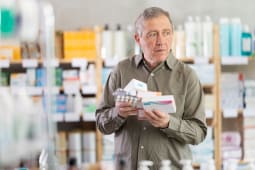 Un homme g dans une pharmacie semble dpass tout en tenant plusieurs botes de pilules et des plaquettes thermoformes, essayant de choisir le bon mdicament.