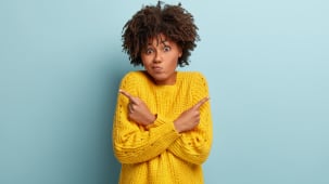 Femme perplexe et confuse ayant une expression hsitante, une coiffure boucle, croisant les mains sur la poitrine, pointant  droite et  gauche sur diffrents cts, portant un pull jaune, tant indiffrente, posant sur un mur bleu