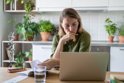 Une femme pensive examine les factures avec un ordinateur portable et un verre d'eau dans la cuisine de sa maison. 
