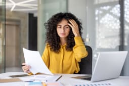 Une femme proccupe examine des documents dans son bureau, apparaissant stresse pendant qu'elle travaille sur son ordinateur portable et analyse des papiers sur son bureau.