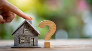 Doigt pointant une maison miniature et un point d'interrogation en bois.