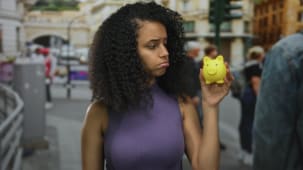 Femme tenant une tirelire dans une rue anim�e, refl�tant une pr�occupation financi�re sur fond urbain, soulignant son identit� hispanique et sa mode d�contract�e au milieu de la foule en plein air.