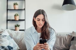 Une jeune femme stress�e essaye d'utiliser internet sur son t�l�phone, rencontrant des probl�mes informatiques chez elle assise sur le canap�, tenant son t�l�phone.