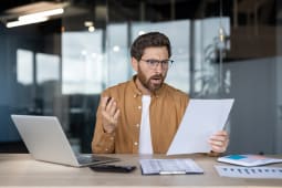 Un hommr assis &agrave; son bureau, tenant des documents et r&eacute;agissant avec une expression choqu&eacute;e.