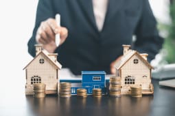 Personne utilisant un stylet derri&egrave;re des maisons miniatures, des pi&egrave;ces empil&eacute;es sur un bureau.