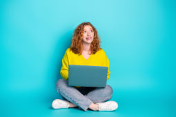Jeune femme aux cheveux rouges assise en tailleur utilisant un ordinateur portable sur un fond turquoise vif, pull d&eacute;contract&eacute;, mode et mode de vie moderne
