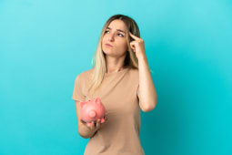 Jeune femme tenant une tirelire sur un fond bleu isol&eacute; ayant des doutes et r&eacute;fl&eacute;chissant