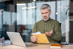 Homme d'affaires mature assis &agrave; son bureau, ouvrant un document d'une enveloppe et r&eacute;agissant avec choc et confusion &agrave; des mauvaises nouvelles inattendues ou &agrave; des probl&egrave;mes financiers.
