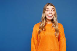 Jeune femme caucasienne choqu&eacute;e, surprise, excit&eacute;e et heureuse, elle porte un pull jaune d&eacute;contract&eacute; regardant de c&ocirc;t&eacute; sur une maquette d'espace de travail isol&eacute;e sur un fond bleu uni en studio. Concept de style de vie.