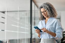 Femme d'affaires d'&acirc;ge moyen heureuse tenant un t&eacute;l&eacute;phone portable et l'utilisant au bureau. Souriant, la femme professionnelle &acirc;g&eacute;e et mature, propri&eacute;taire d'une entreprise, utilise son smartphone debout dans le couloir.