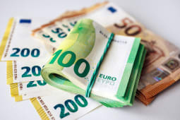 Pile de billets en euros comprenant un rouleau de billets de 100 euros sur une surface blanche avec des d&eacute;nominations suppl&eacute;mentaires de 50 et 200 euros visibles en arri&egrave;re-plan