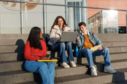 Trois &eacute;tudiants assis sur des marches ext&eacute;rieures, utilisant un ordinateur portable et discutant.