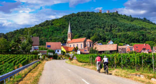 Deux cyclistes traversent un village entour&eacute; de vignes verdoyantes. Couple cycling on Alsace Wine Route through vineyards to Husseren-les-Ch&acirc;teaux village, France Active slow travel lifestyle.