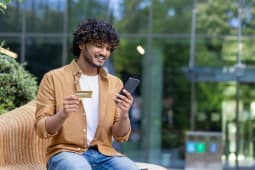 Un jeune indien occup&eacute; est assis dehors sur un banc, tenant une carte de cr&eacute;dit et utilisant un t&eacute;l&eacute;phone portable