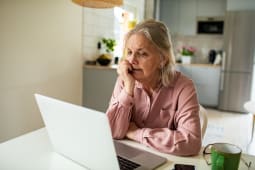 Une femme &acirc;g&eacute;e inqui&egrave;te devant son ordinateur portable dans sa cuisine.