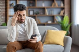 Homme frustr&eacute; assis sur un canap&eacute; v&eacute;rifiant son smartphone, se sentant stress&eacute; &agrave; la maison, portant des v&ecirc;tements d&eacute;contract&eacute;s