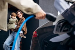 IUne femme debout portant un enfant pendant qu'une autre personne charge une voiture &eacute;lectrique.