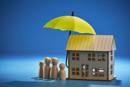Des figurines miniatures d'une famille se tiennent &agrave; c&ocirc;t&eacute; d'une maisonsur laquelle est plac&eacute;e un parapluie pour symboliser l'assurance.