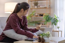 Une jeune femme de bonne humeur assise sur le canap&eacute; dans le salon et utilisant un ordinateur portable et une carte de cr&eacute;dit.