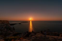Des gens regardent une &eacute;clipse solaire totale au coucher du soleil depuis la c&ocirc;te rocheuse de Minorque avec un reflet d'&eacute;clat solaire sur l'oc&eacute;an