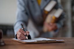 Main d'affaires tenant un stylo et signant un contrat plac&eacute; sur un presse-papiers, tout en portant &eacute;galement des livres de droit, symbolisant l'expertise juridique, l'accord et les documents officiels.