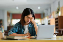 Jeune femme stress&eacute;e en &eacute;tudiant avec un ordinateur portable &agrave; la biblioth&egrave;que, &eacute;tudiante d&eacute;bord&eacute;e se pr&eacute;parant pour les examens, pression acad&eacute;mique et sant&eacute; mentale