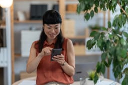 Femme asiatique petite entrepreneuse v&eacute;rifiant son smartphone dans un bureau &agrave; domicile lumineux, souriant tout en g&eacute;rant ses t&acirc;ches commerciales avec concentration et &eacute;nergie