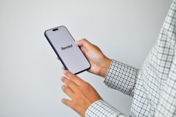 Personne en chemise &agrave; carreaux tenant un smartphone moderne affichant l'application de banque num&eacute;rique Revolut sur un &eacute;cran blanc vif sur fond gris clair