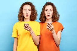 Jumeaux &eacute;motionnels surpris avec la bouche grande ouverte et les yeux &eacute;carquill&eacute;s tenant des smartphones et regardant la cam&eacute;ra. Vente, commerce, message gratuit, doubles r&eacute;ductions