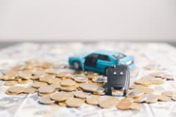 Cl&eacute; de voiture entour&eacute;e de pi&egrave;ces et de billets avec un v&eacute;hicule jouet en arri&egrave;re-plan.