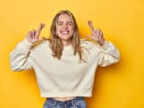 Jeune femme blonde en sweatshirt blanc sur un fond de studio jaune, croisant les doigts pour avoir de la chance
