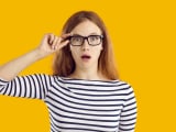 Jeune étudiante très surprise par quelque chose. Belle jeune femme nerd en haut rayé et lunettes isolée sur fond jaune regardant la caméra avec une expression faciale drôle, choquée, étonnée