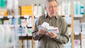 Un homme âgé dans une pharmacie semble dépassé tout en tenant plusieurs boîtes de pilules et des plaquettes thermoformées, essayant de choisir le bon médicament.
