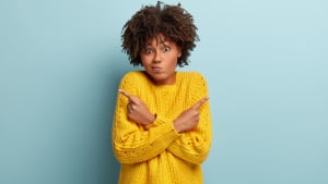 Femme perplexe et confuse ayant une expression hésitante, une coiffure bouclée, croisant les mains sur la poitrine, pointant à droite et à gauche sur différents côtés, portant un pull jaune, étant indifférente, posant sur un mur bleu