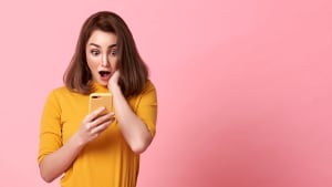 femme jeune, heureuse et surprise, belle à regarder, utilise un téléphone portable sur un fond rose avec de l'espace pour du texte.
