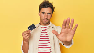 Un homme tenant une carte de crédit sur fond jaune se tient la main tendue montrant un signe d'arrêt, vous empêchant.