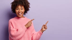 Plan d'une femme charmante et positive, qui montre quelque chose dans un espace vide, a une expression de joie, porte un pull rose ample, fait des gestes sur un mur violet.