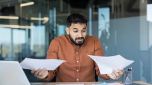 Homme d'affaires confus tenant deux documents, comparant les données et les rapports tout en ressentant du stress et de l'incertitude concernant les informations financières et la paperasse au bureau