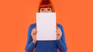Jeune femme en pull bleu tenant une feuille de papier vierge devant son visage contre un fond orange vif lors d'une séance photo créative