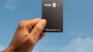 Main tenant une carte bancaire Visa Boursobank devant ciel bleu.