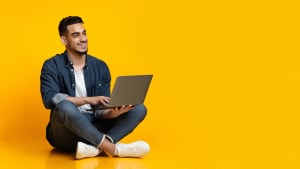 Jeune homme arabe heureux assis sur le sol, utilisant un nouvel ordinateur portable moderne, tapant sur le clavier et souriant sur fond de studio jaune, regardant l'espace de copie pour le texte ou la publicité, panorama
