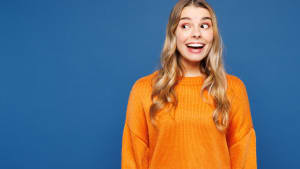 Jeune femme caucasienne choqu�e, surprise, excit�e et heureuse, elle porte un pull jaune d�contract� regardant de c�t� sur une maquette d'espace de travail isol�e sur un fond bleu uni en studio. Concept de style de vie.