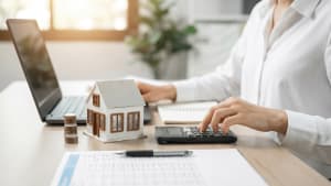 Personne calcule finances immobilières avec ordinateur, maison miniature, pièces.