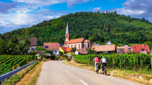 Deux cyclistes traversent un village entour� de vignes verdoyantes. Couple cycling on Alsace Wine Route through vineyards to Husseren-les-Ch�teaux village, France Active slow travel lifestyle.