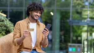 Un jeune indien occup� est assis dehors sur un banc, tenant une carte de cr�dit et utilisant un t�l�phone portable