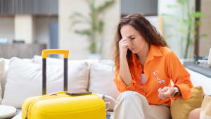 Une femme stressée tenant des lunettes et touchant son visage tout en étant assise près d'une valise.