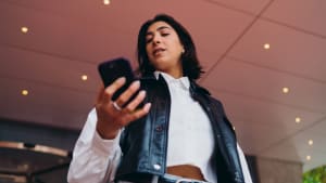 Étudiante espagnole vérifiant les horaires de transport sur une application smartphone. Femme hispanique envoyant des messages texte sur l'écran de son téléphone mobile près d'un pavillon de construction architecturale moderne dans un couloir.