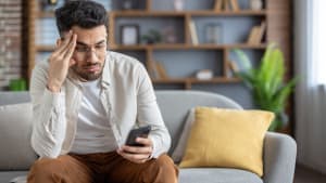 Homme frustré assis sur un canapé vérifiant son smartphone, se sentant stressé à la maison, portant des vêtements décontractés