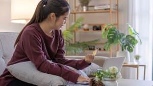 Une jeune femme de bonne humeur assise sur le canapé dans le salon et utilisant un ordinateur portable et une carte de crédit.