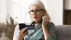 Femme âgée inquiète et surprise en lunettes regardant un smartphone et une carte de crédit, rencontrant des problèmes financiers, mauvais service bancaire en ligne, découvrant des dépenses excessives, risque de faillite